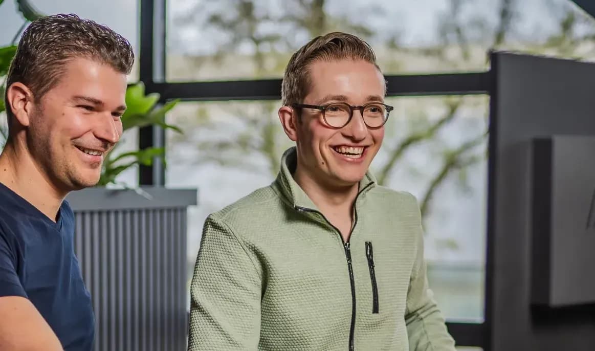 Twee lachende mannen kijken naar een computerscherm in een lichte ruimte met raam