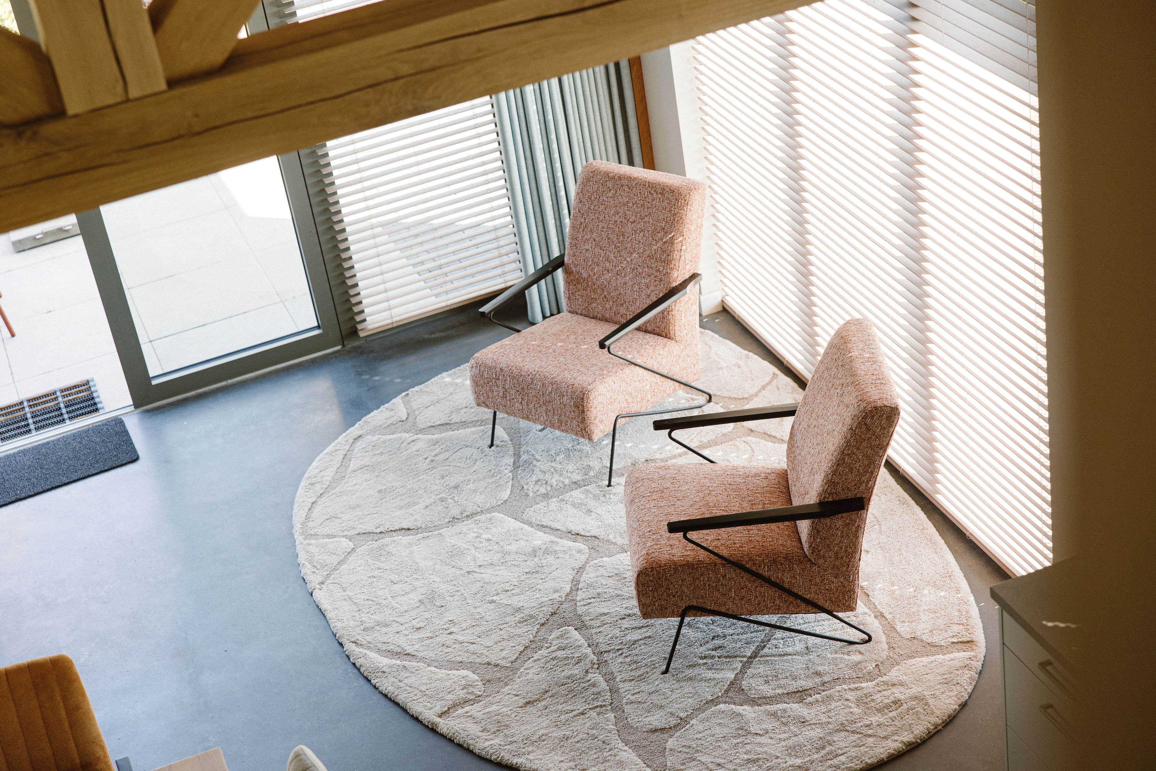 Bovenaanzicht van twee gestoffeerde stoelen op een rond beige vloerkleed naast grote ramen met witte jaloezieën