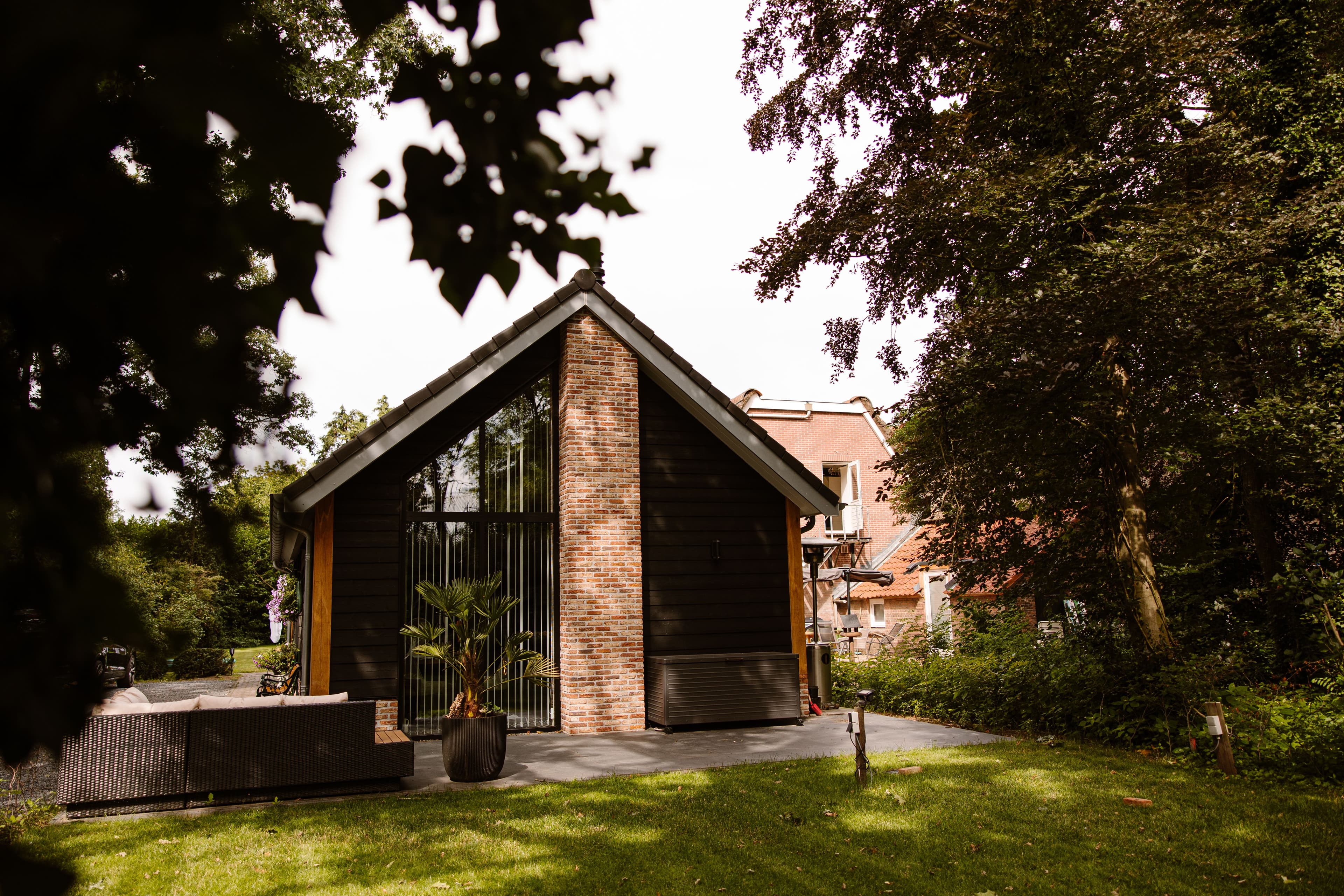 Vooraanzicht van een gebouw met bakstenen schoorsteen, donkere houten gevel en grote glazen pui; op het terras staat een rieten loungebank en een potplant, rondom bomen.