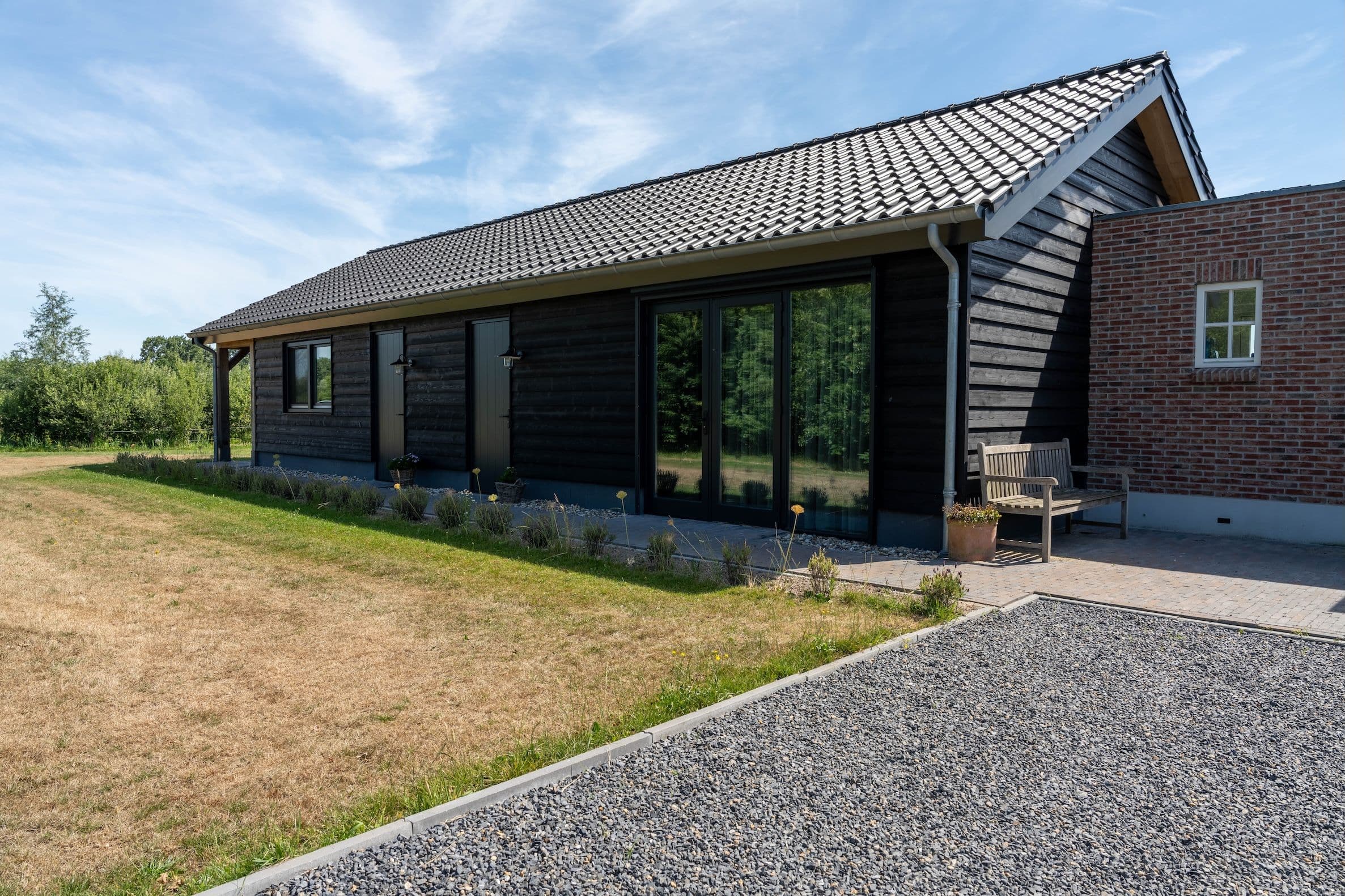 Zijaanzicht van een huis met zwarte houten gevel, glazen schuifpui, bakstenen aanbouw, donkergrijze dakpannen en houten bank op het terras