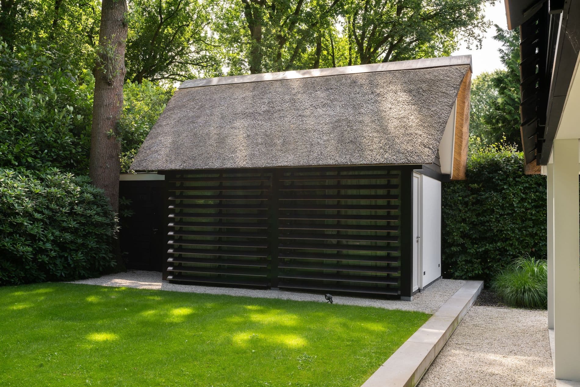 Tuinhuis met rieten dak en zwarte lamellengevel voor een strak onderhouden gazon, omgeven door bomen en hagen.